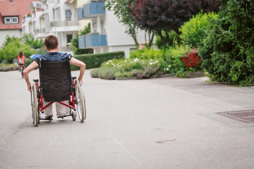 Frau fährt Rollstuhl an Straße Rückenansicht 20160725-0477 