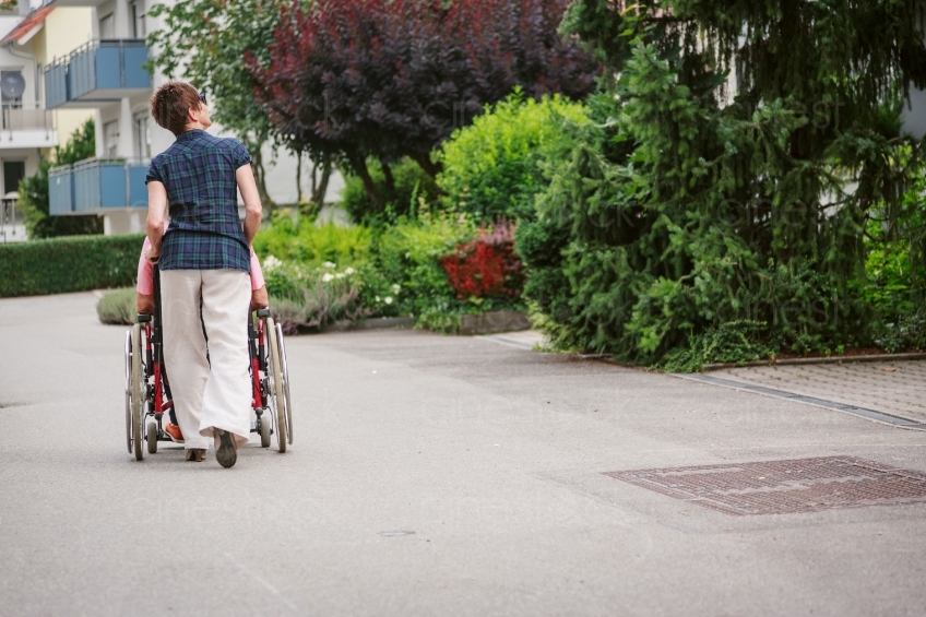 Frau schiebt Rollstuhl an Straße Rückenansicht 20160725-0430 