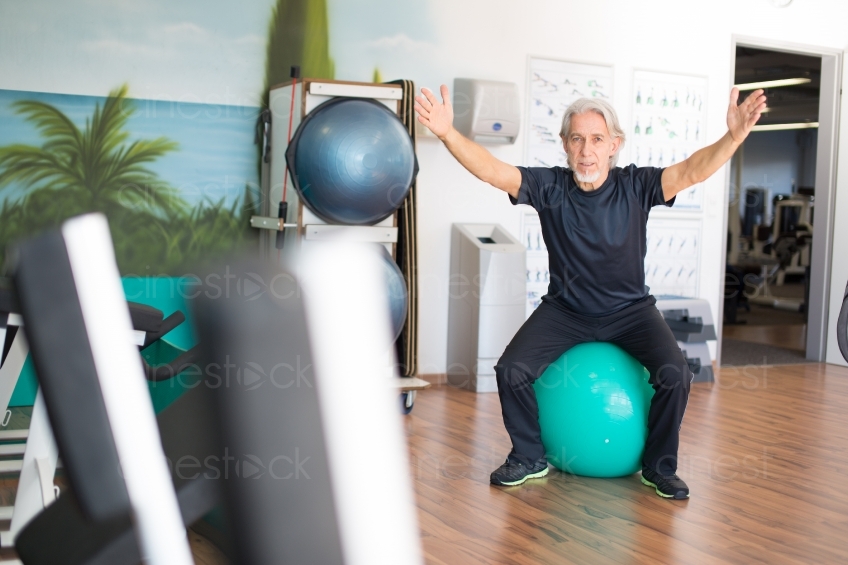 Älterer Mann macht Übungen auf Gymnastikball mit gestreckten Armen 20160212-0311