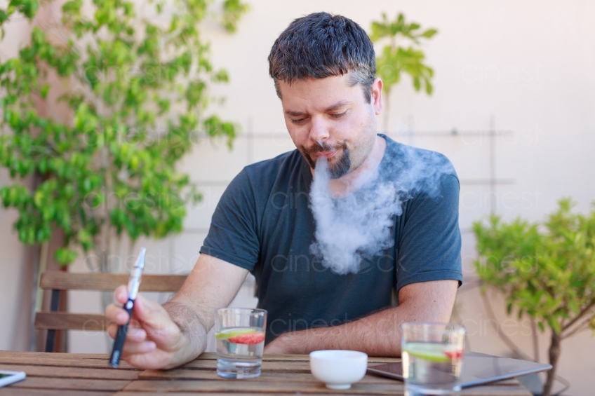 Mann sitzt mit E-Zigarette und Getränken an einem Tisch 20160823