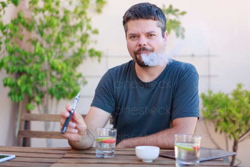 Mann sitzt mit E-Zigarette und Getränken an einem Tisch 20160823